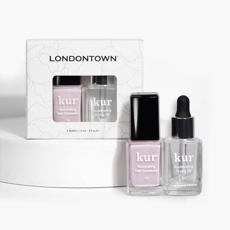 LONDONTOWN's "Perfect in a Pinch: Pink" nail care set features the kur illuminating nail concealer and accelerator drying oil in clear bottles. One bottle includes a brush applicator, while the other comes with a dropper. The elegant packaging in the background enhances this kit designed for polished perfection.