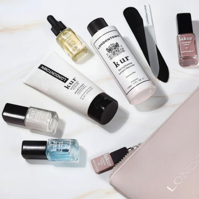 A flat lay of the LONDONTOWN Discovery Set 2.0 displays nail care essentials—polishes, lotion, cuticle oil, a nail file, and a beige zippered pouch—arranged on a light surface.