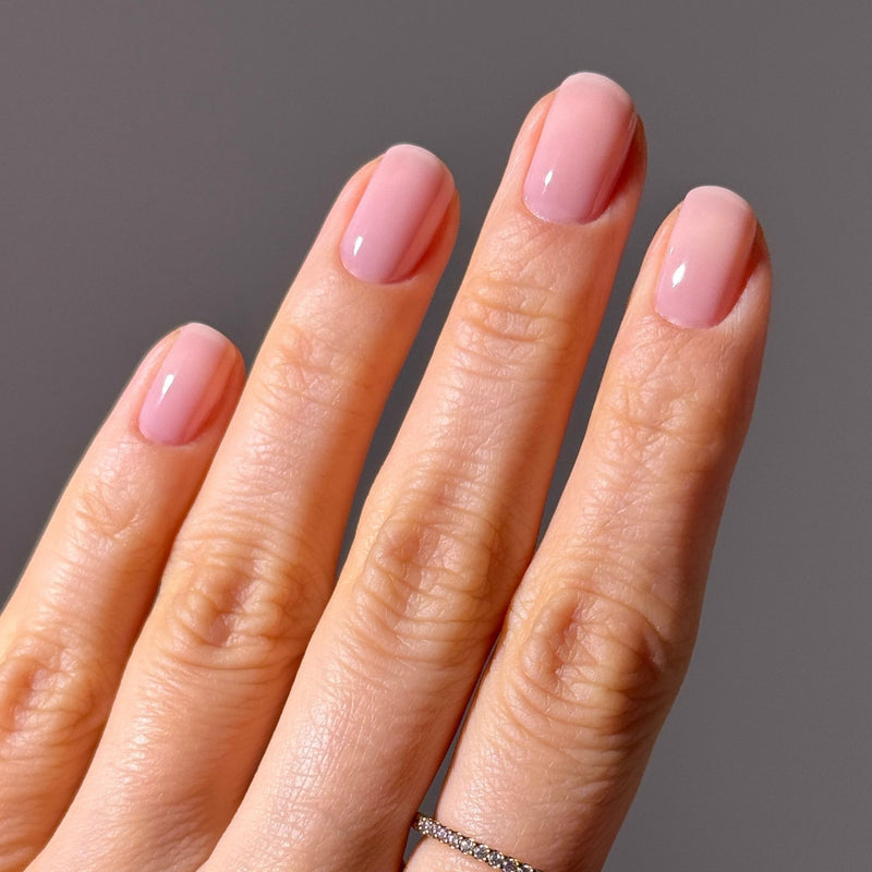 A hand with neatly manicured nails painted in LONDONTOWN’s Dream State, a demi-sheer cream pink, rests against a plain gray background. One finger is adorned with a thin, delicate ring.