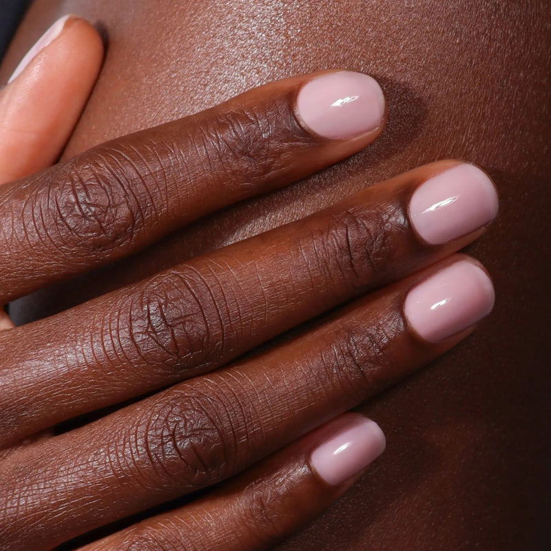 A close-up of a hand with short, neatly manicured nails painted in LONDONTOWN’s Dream State, a demi-sheer cream pale pink, resting on dark brown skin and giving off a soft glow.