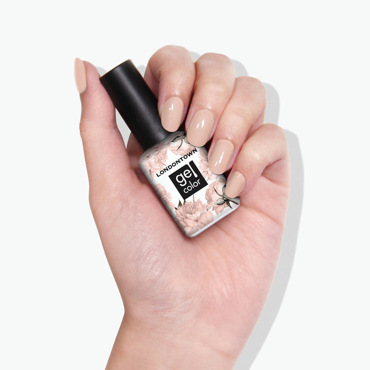 A hand with a nude manicure holds a bottle of LONDONTOWN Dusk gel color nail polish, showcasing an easy-to-wear, neutral peach shade.