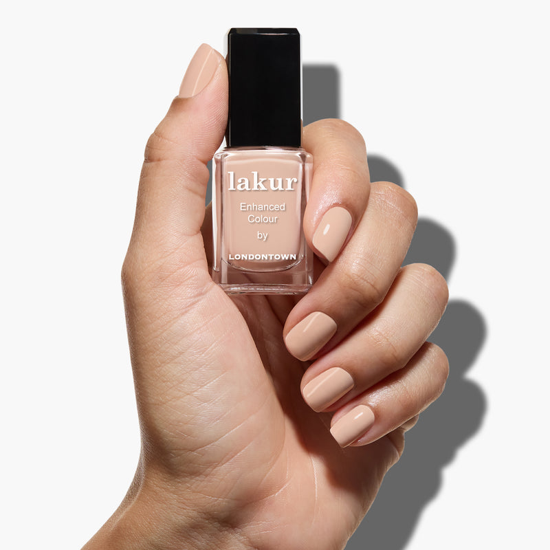 A hand with manicured nails holds a bottle of LONDONTOWN Dusk nail polish in a neutral, nude shade against a white background.