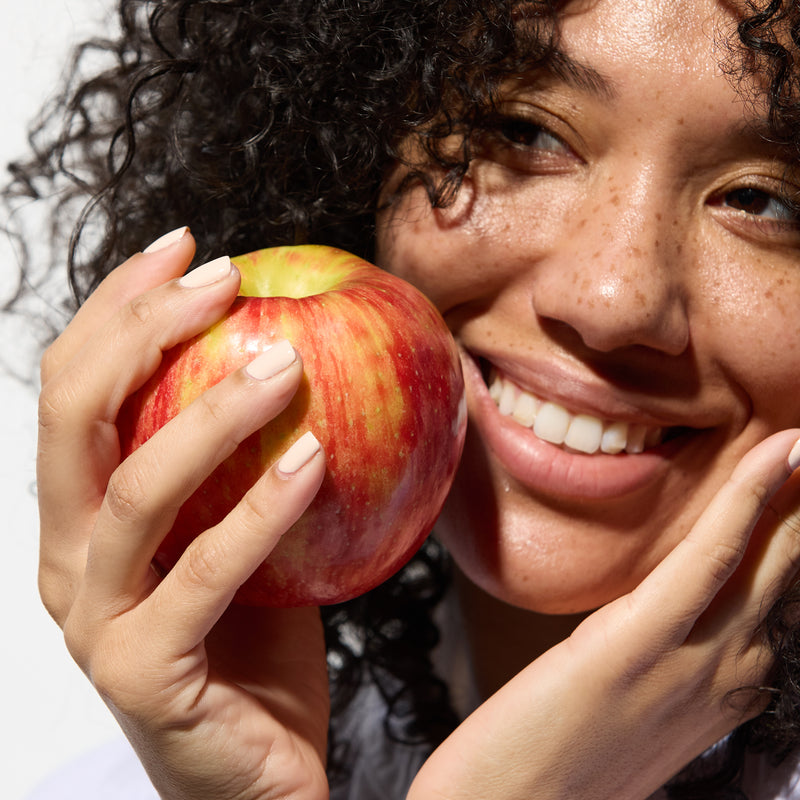 A person with curly hair smiles while holding a peach close to their face, showcasing the peach and their white nails polished with Dusk by LONDONTOWN.