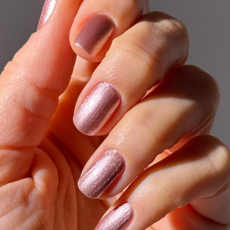 A close-up of a hand with neatly manicured nails painted in LONDONTOWN's Extra Bubbles, Please—a shimmering mauve rose gold polish.