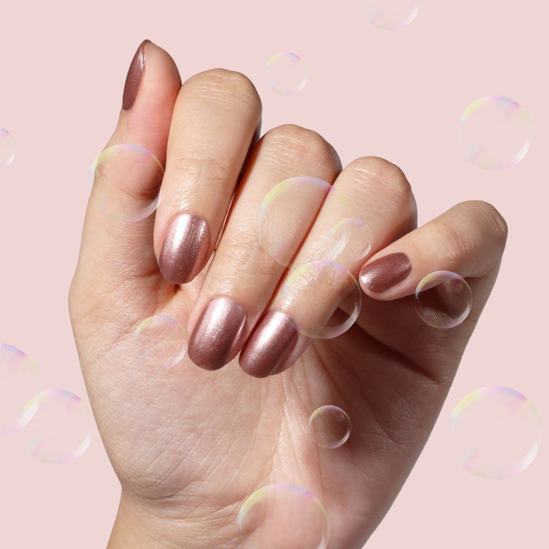 A hand with neatly manicured nails painted in LONDONTOWN’s Extra Bubbles, Please—a shimmering mauve gold polish—is shown against a light pink background with translucent bubbles.