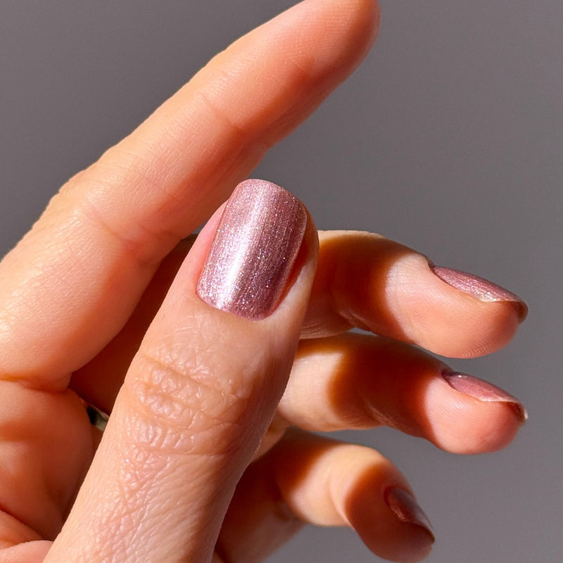 A hand with short, well-groomed nails painted in LONDONTOWN's Extra Bubbles, Please shimmery mauve gold nail polish glows softly against a plain background.