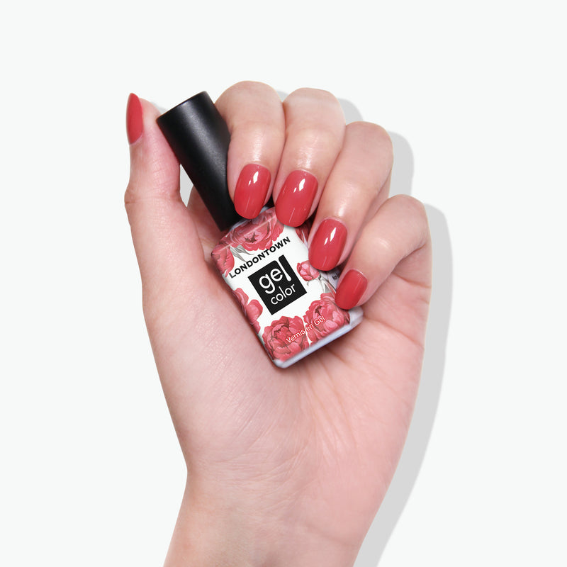 A hand with red-painted nails holds a bottle of LONDONTOWN's Flushed Cheeks nail polish, the sun-kissed hue adding a touch of warmth.
