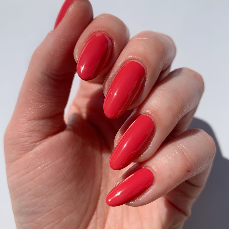 A hand with neatly manicured nails, each painted in LONDONTOWN's glossy red shade, Flushed Cheeks, held up against a plain background.