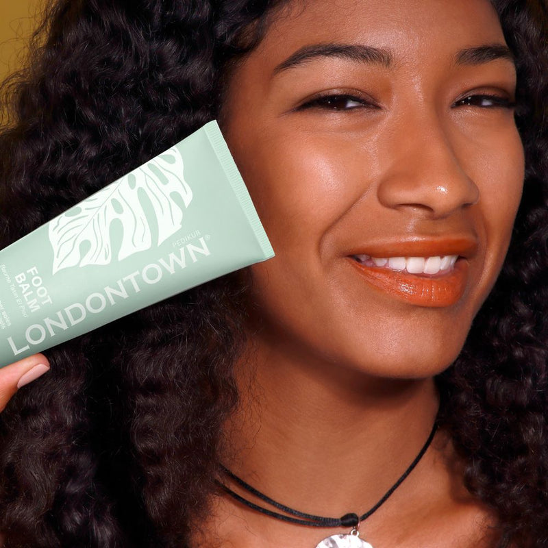 A person smiling while holding a green tube of LONDONTOWN Foot Balm with eucalyptus.