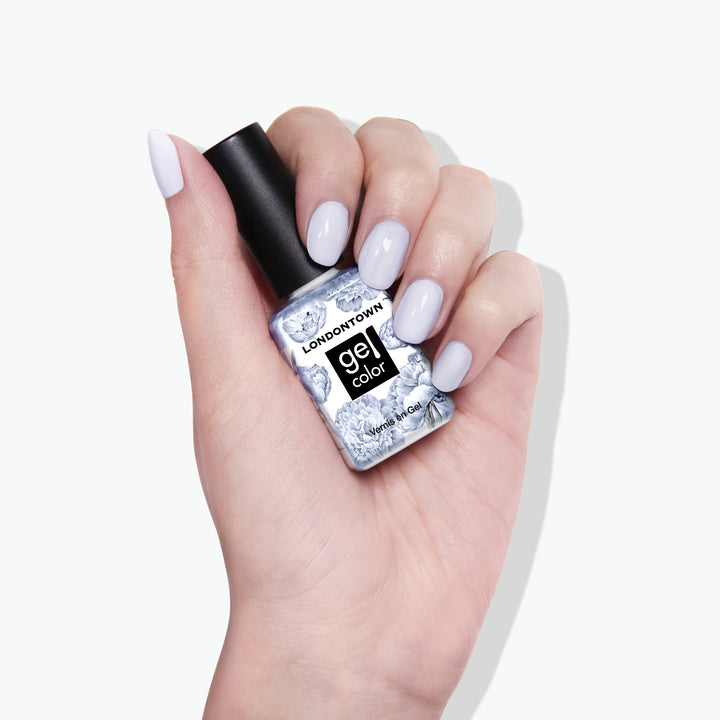A hand with light purple nail polish holds a LONDONTOWN Frostbitten Gel Color nail polish bottle with a black cap and marbled gray and white label under bright lighting, hinting at shades of ice blue in the background.