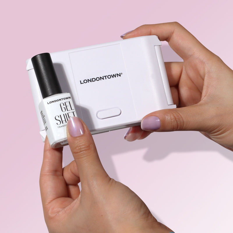 A person holds a white LONDONTOWN GelShift Kit UV nail lamp in one hand and a LONDONTOWN GelShift Kit gel polish bottle in the other, ready for a nail transformation against a light pink background.