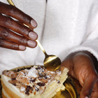 A person in a white robe enjoys LONDONTOWN's "Champagne in Bed" cake, using a gold spoon to savor it. Its neutral pink frosting glistens, with the crumb topping adding an inviting touch.