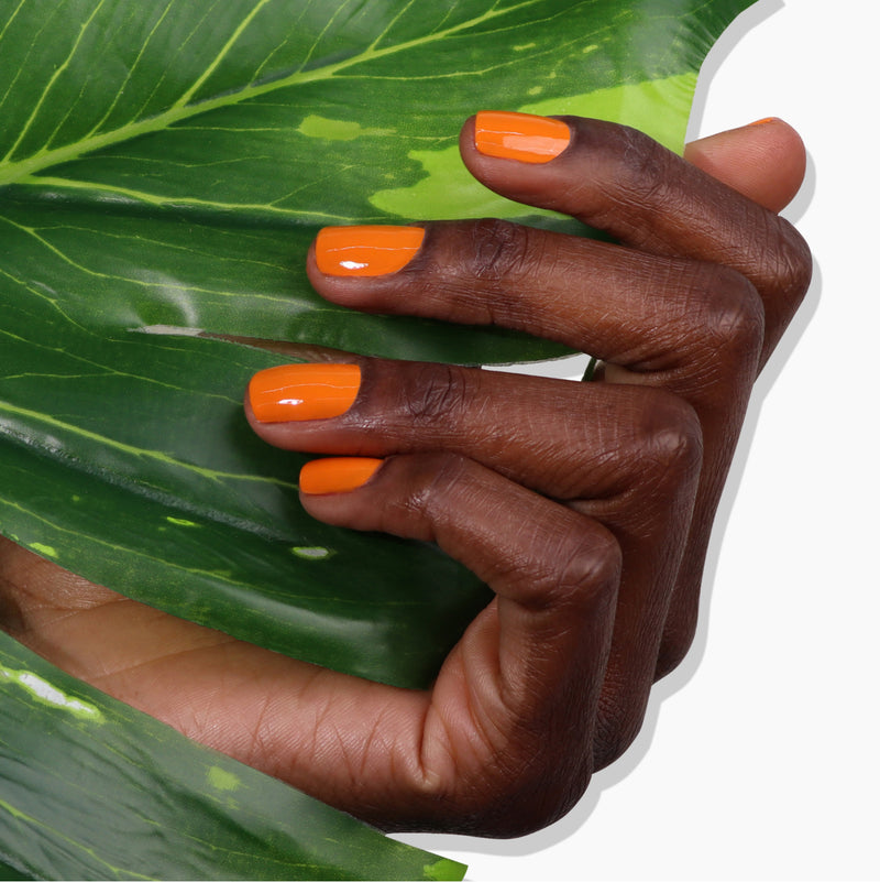 A hand with LONDONTOWN's vibrant burnt orange "Heat Wave" nail polish partially concealed by large green leaves.