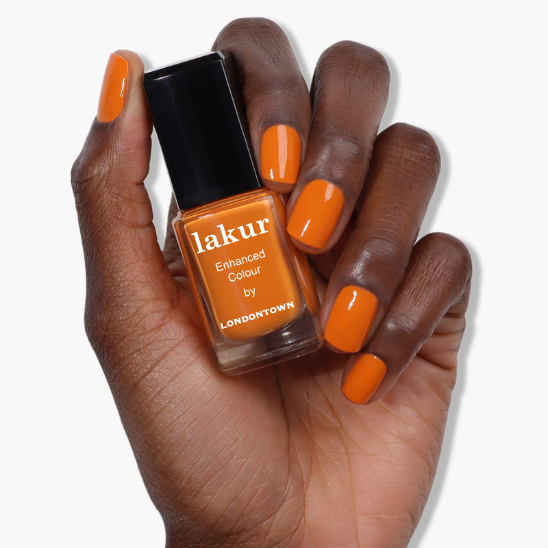 A hand with a dark skin tone holds a bottle of burnt orange nail polish labeled "Heat Wave by LONDONTOWN." The nails are painted the same vibrant shade of orange.