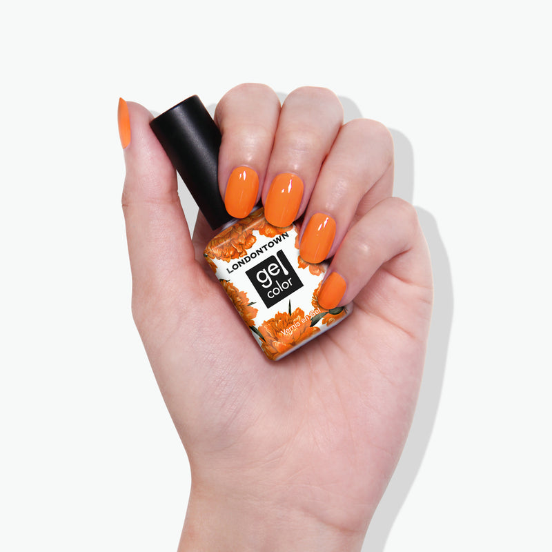 A hand with burnt orange-painted nails holds a bottle of LONDONTOWN Heat Wave Gel Color nail polish.