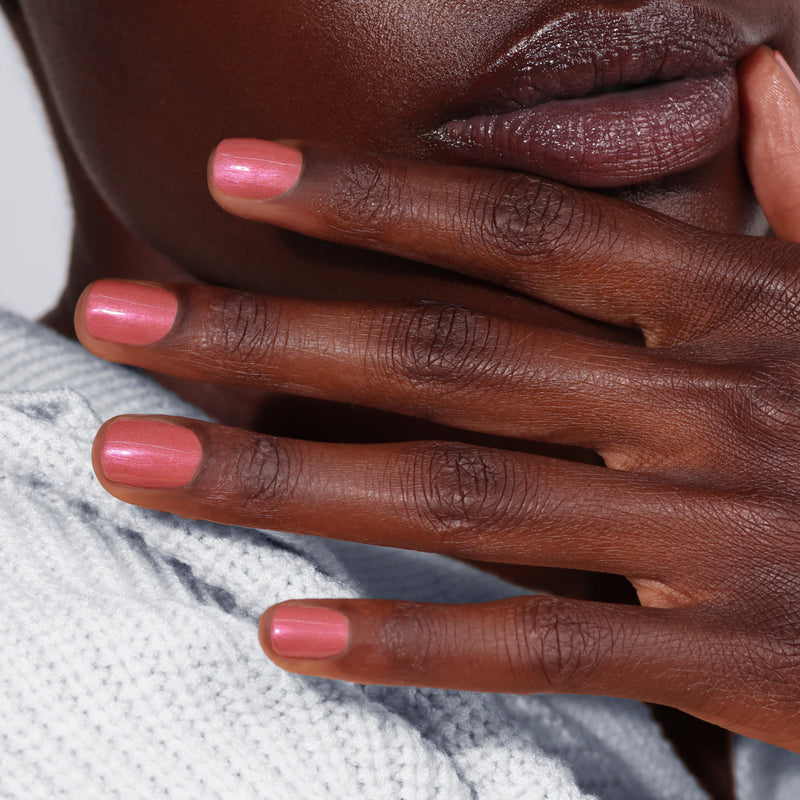 Close-up of a hand adorned with LONDONTOWN's Slopeside pink chrome nail polish resting against a face with natural lips, set against a textured white fabric.