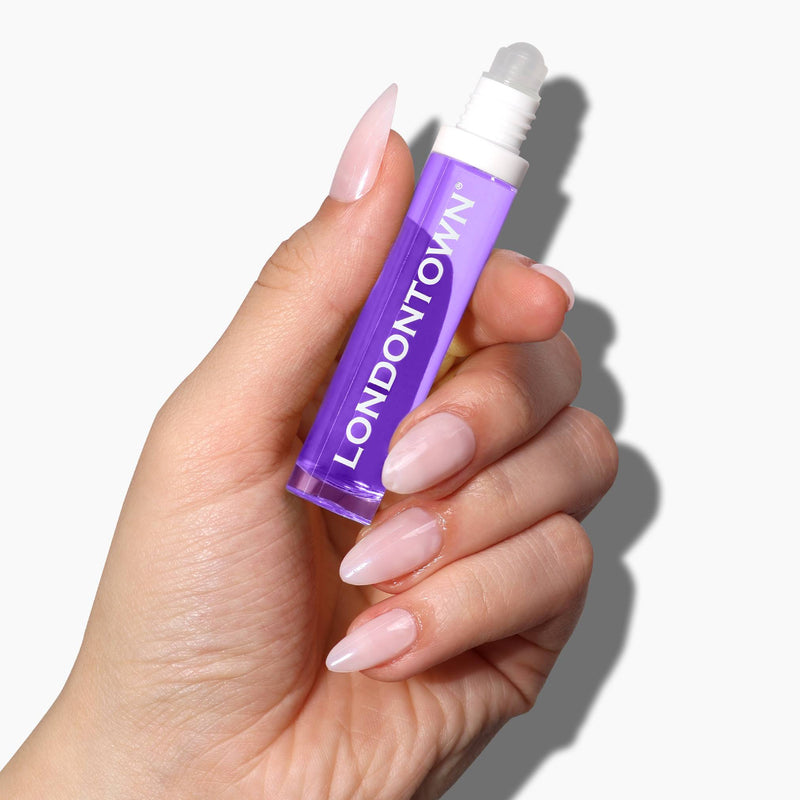A hand holding the LONDONTOWN Nighttime Cuticle Quench - Lavender, which has a purple liquid inside, set against a white background, promising soothing cuticle treatment.