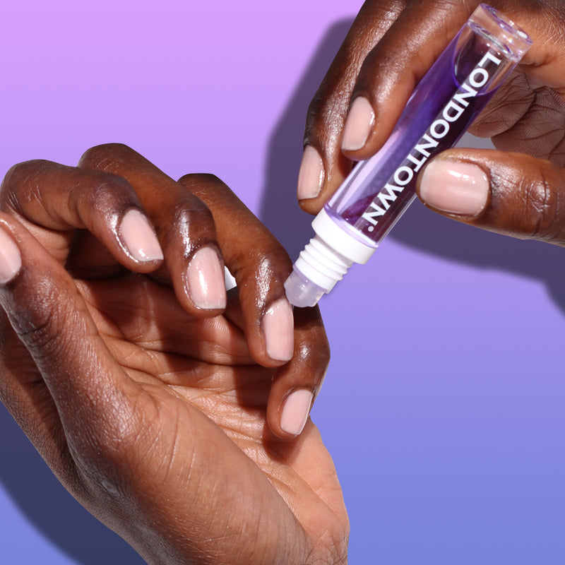 Close-up of hands applying Nighttime Cuticle Quench - Lavender from a tube labeled "LONDONTOWN" onto fingernails, set against a gradient purple background, offering soothing cuticle treatment.