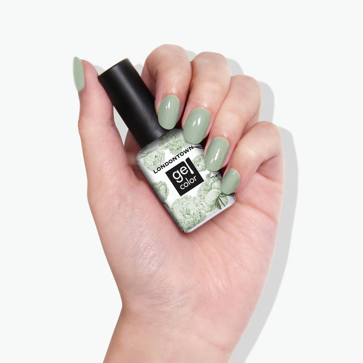 A hand with painted fingernails holds a bottle of LONDONTOWN gel color nail polish in the shade "Lichen," a light green reminiscent of white sage.