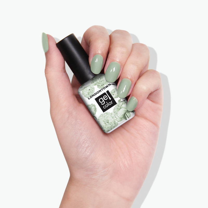 A hand with painted fingernails holds a bottle of LONDONTOWN gel color nail polish in the shade "Lichen," a light green reminiscent of white sage.