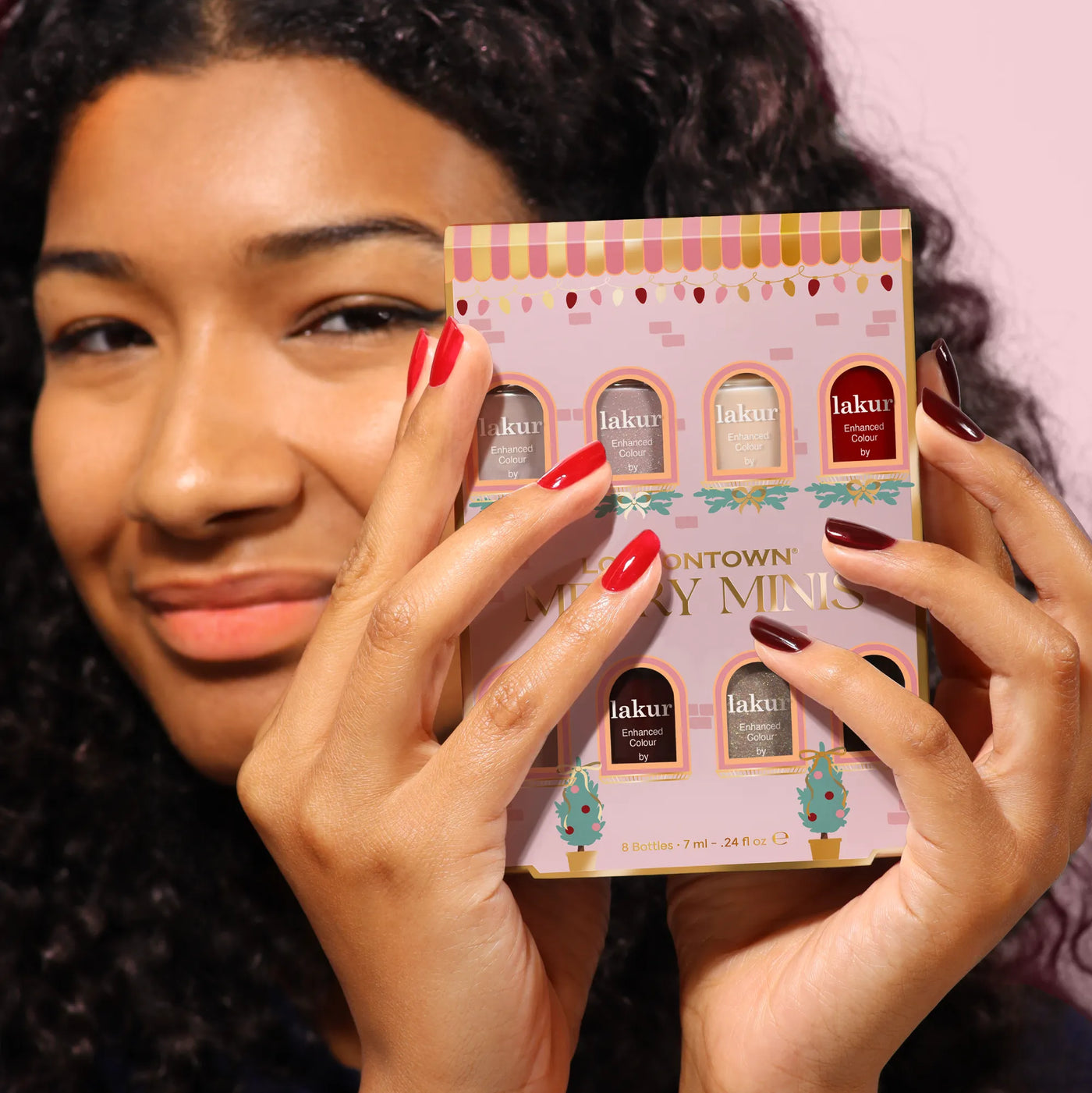 A person holds up the LONDONTOWN Merry Minis Set, showcasing six festive nail polish shades perfect for holiday manis, with their nails painted in vibrant red and deep burgundy.