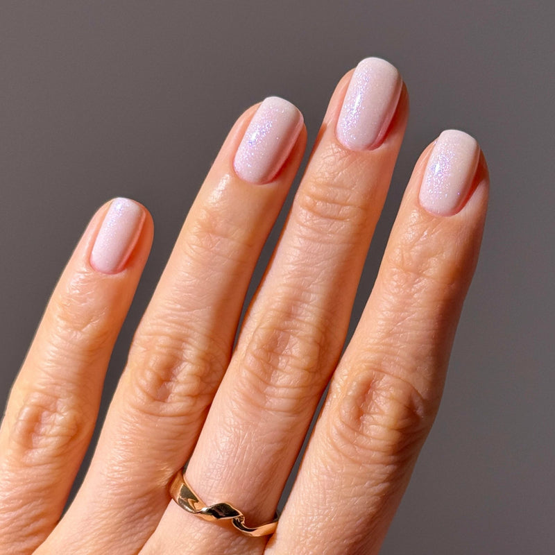 A hand with a gold ring displays neatly manicured nails painted in Milk Bath by LONDONTOWN, a demi-sheer cream polish with a subtle soap bubble iridescence.