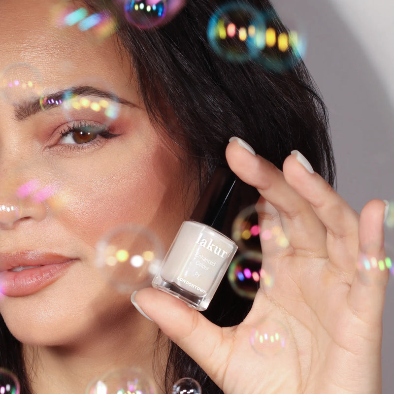 A person holds a bottle of LONDONTOWN Milk Bath demi-sheer cream nail color near their face, surrounded by shimmering bubbles with soap bubble iridescence.