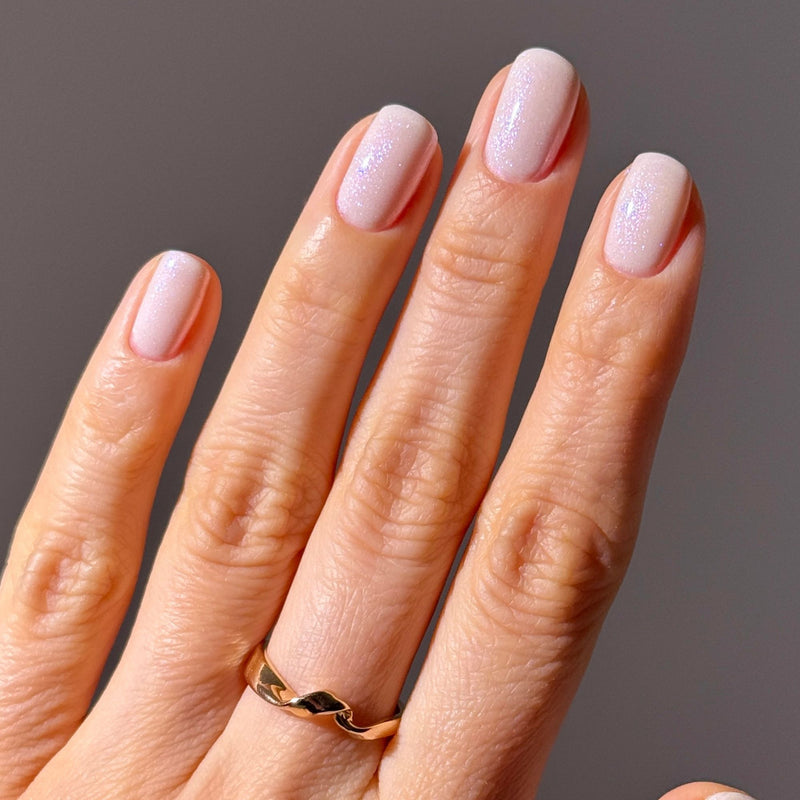 A hand with neatly manicured nails painted in LONDONTOWN's Milk Bath, a light pink iridescent cream polish, wearing a gold ring on the ring finger against a neutral background.