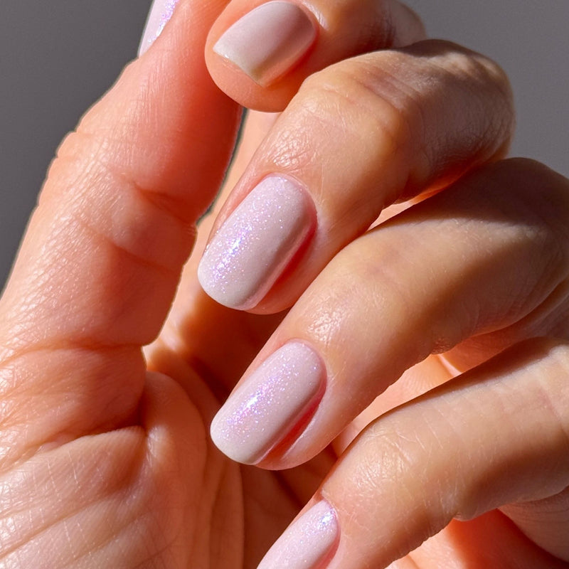 A hand with neatly manicured nails is painted in LONDONTOWN's Milk Bath, a pale pink demi-sheer cream polish with subtle shimmer, shown in natural light.