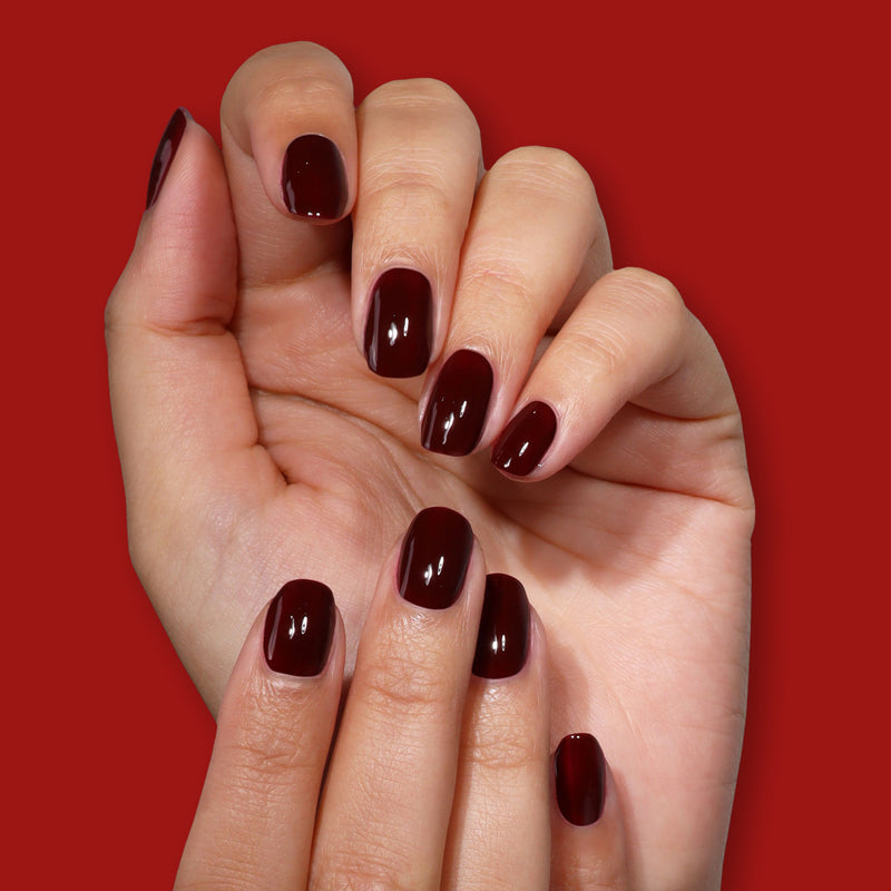 A hand with neatly manicured nails painted in LONDONTOWN's Naughty List, a glossy merlot red, is displayed against a matching solid red background.