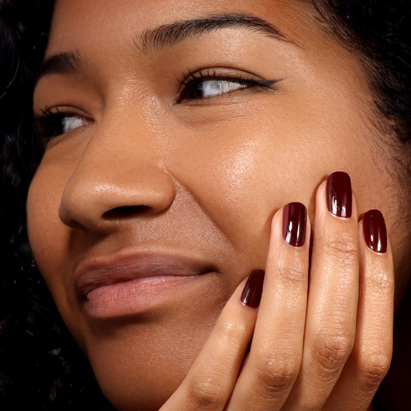 A person with smooth skin shows off their cheek and hand with LONDONTOWN's Naughty List—a dark red, full-bodied merlot nail polish—applied to their nails in a close-up shot.