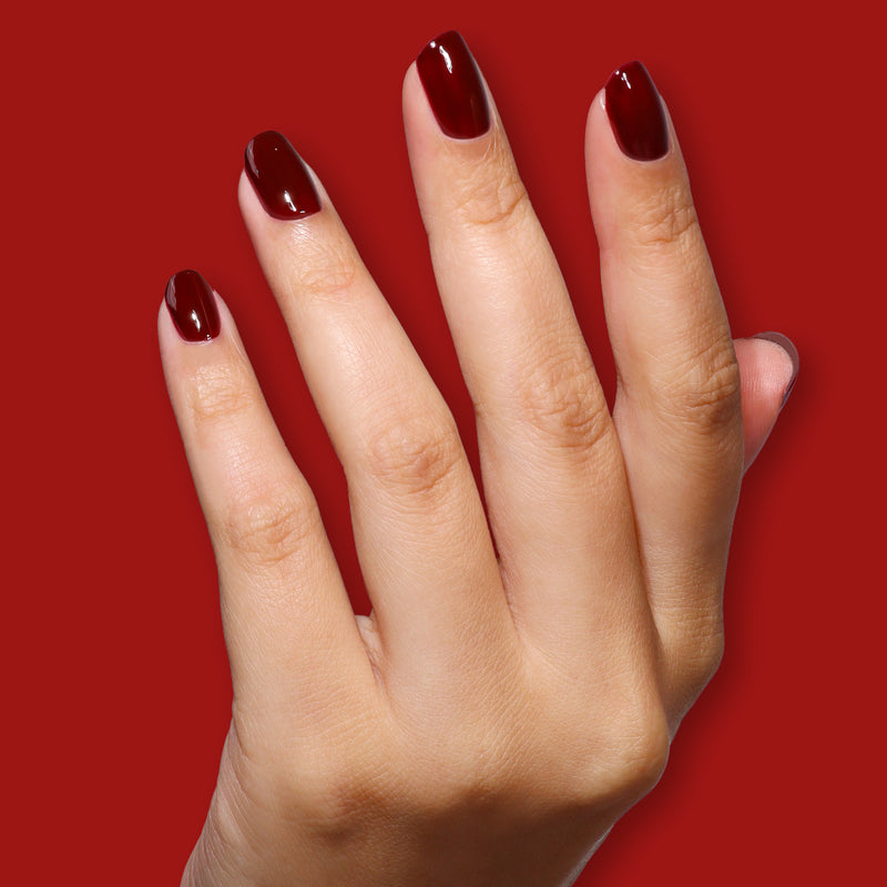 A hand with neatly manicured nails painted in LONDONTOWN’s Naughty List, a rich merlot red, is shown against a solid red background.