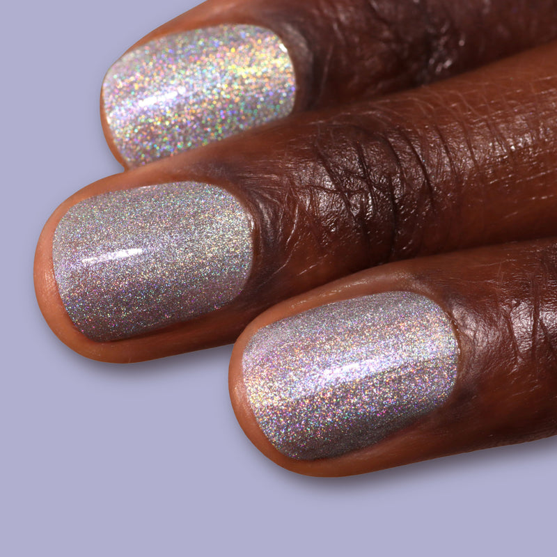 Close-up of three dark-skinned fingers with nails painted in LONDONTOWN's Nice & Icy, a shiny platinum holographic silver polish, set against a light purple background.