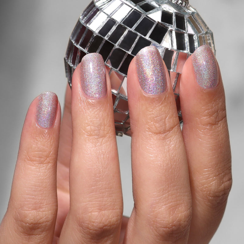 A hand with short, oval nails painted in LONDONTOWN’s Nice & Icy platinum holographic polish holds a small silver disco ball.