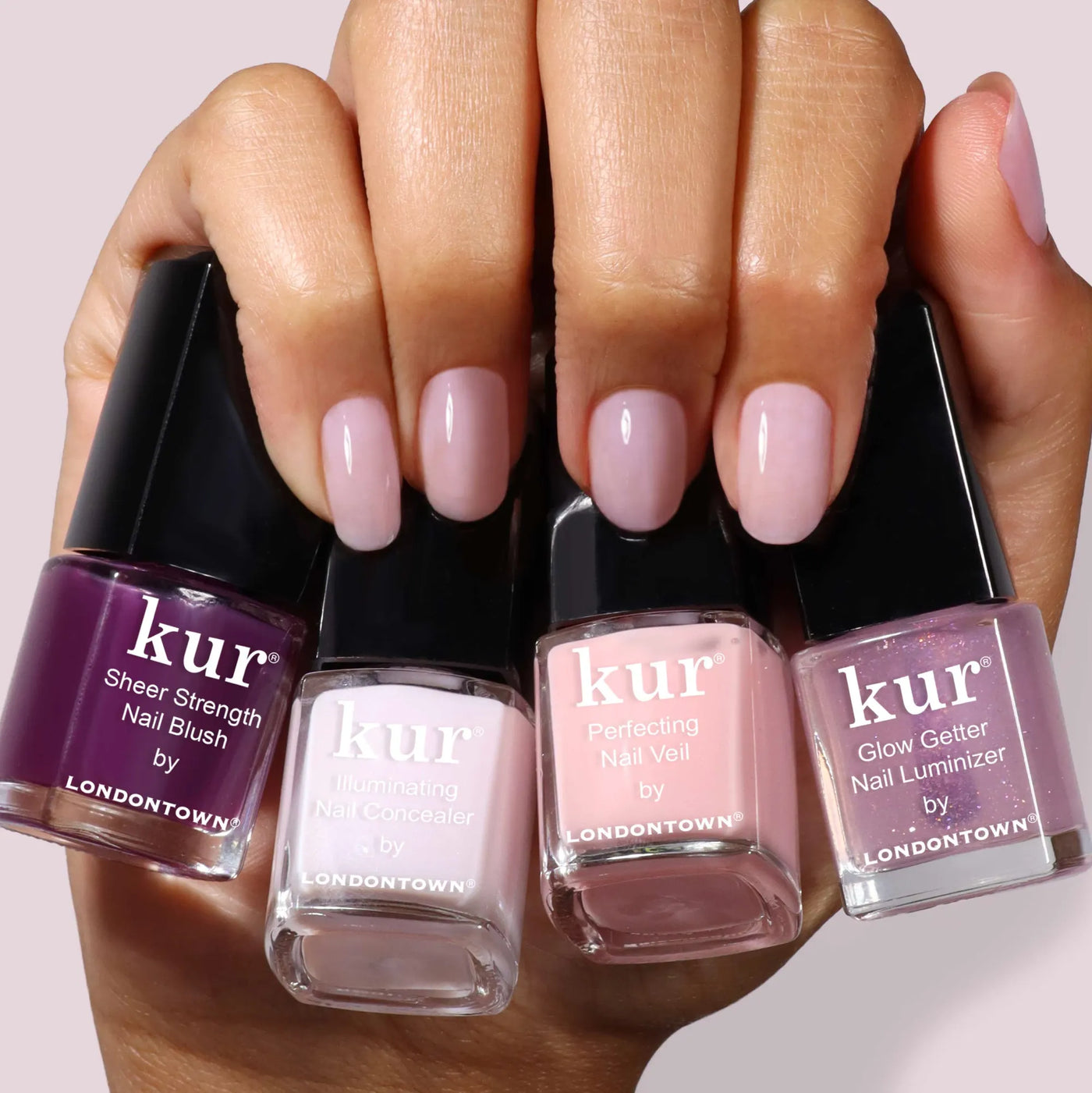 A hand with manicured nails holds four bottles of LONDONTOWN One-Step Hero Minis in assorted pink and purple shades, combining color and care for a flawless finish.