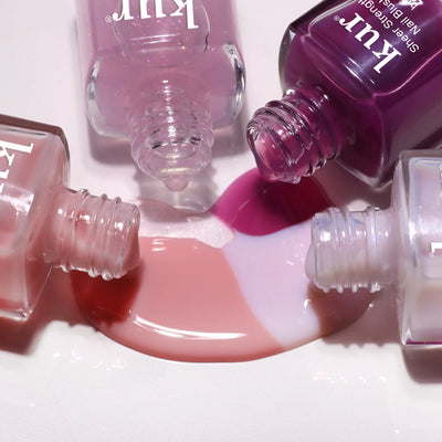 Four open bottles of pink and purple nail polish spill onto a white surface, their colors blending—just as LONDONTOWN’s One-Step Hero Minis deliver effortless color and care in one easy step.