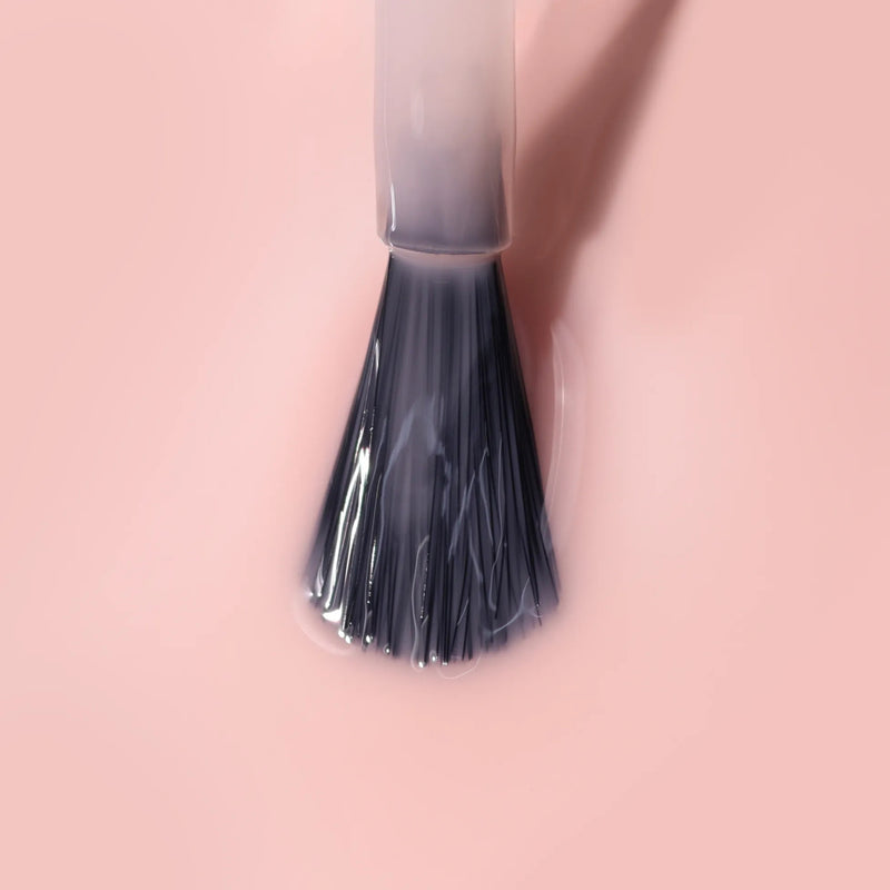 A close-up of LONDONTOWN's Perfecting Nail Veil #4 brush coated with light pink polish, delivering a hint of color and care against a matching pink background.