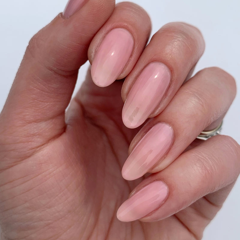 A hand with neatly manicured, almond-shaped nails painted in LONDONTOWN Perfecting Nail Veil #7—a glossy, light pink shade that gives your nails the fresh, moisturizing look of a tinted lip balm.