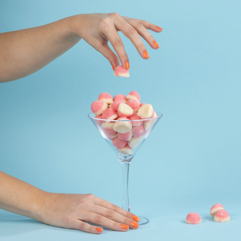Two hands, adorned with the vibrant Paloma orange nail polish by LONDONTOWN, hold and grab pink sugar-coated candy marshmallows from a martini glass against a blue background.