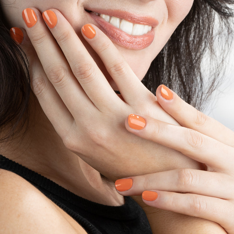 A close-up of a person smiling with their hands touching their face, showcasing shiny fingernails painted with LONDONTOWN's vibrant peach-colored polish, Paloma.
