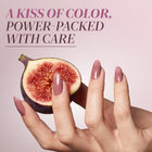 A hand with nails painted in LONDONTOWN’s Perfecting Nail Veil #11 holds half a fig against a pink background. Text above reads: "A KISS OF COLOR, POWER-PACKED WITH CARE." Experience the nourishing touch of care in every power-packed swipe—a hint of color that speaks volumes.