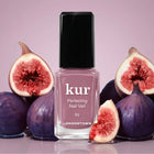 A bottle of LONDONTOWN Perfecting Nail Veil #11 sits surrounded by whole and halved figs against a light purple background, evoking the same fresh elegance as your favorite tinted lip balm or nourishing lip care essential.