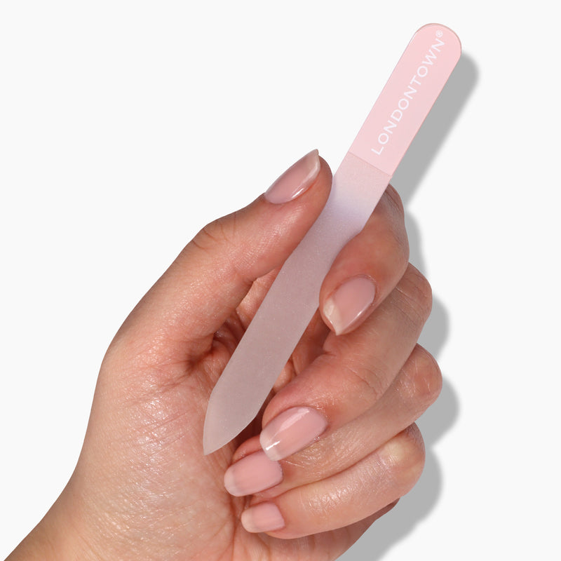 A hand holding a LONDONTOWN Glass Nail File - Petal, featuring a pink and transparent design. The nails are well-manicured and polished with a light, natural color, showcasing effortless nail filing for a damage-free finish.