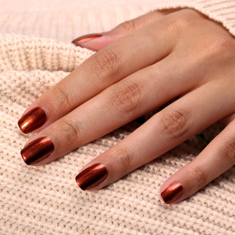 A hand adorned with LONDONTOWN's Posh Forever metallic bronze nails rests on a beige textured fabric, reminiscent of warm cinnamon tones.