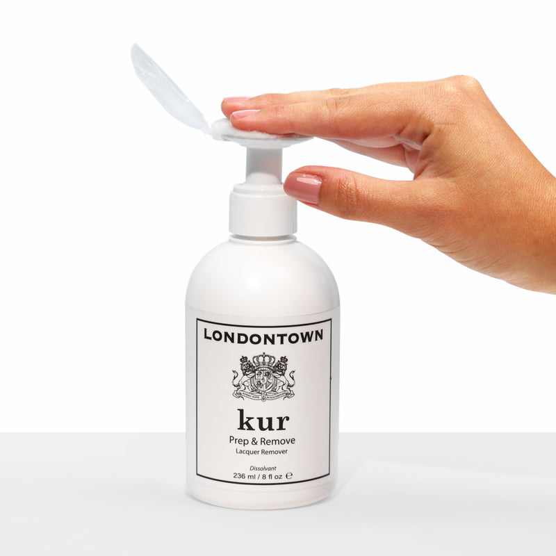 A hand with painted nails presses the pump of a white LONDONTOWN Prep & Remove Lacquer Remover bottle against a plain white background.