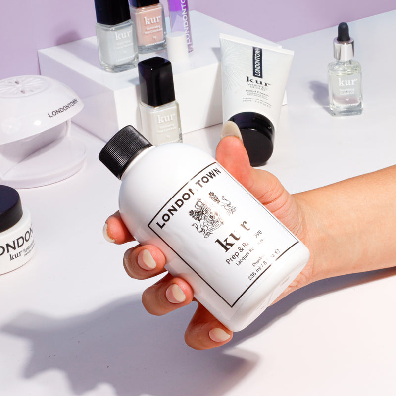 A hand holds a bottle of LONDONTOWN Prep & Remove Lacquer Remover, surrounded by other LONDONTOWN nail care products on a white surface.