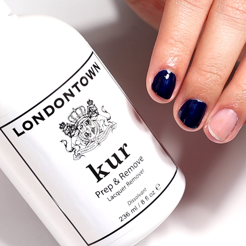 A hand with partial dark nail polish on two nails rests beside a bottle of LONDONTOWN Prep & Remove Lacquer Remover, ready to gently and effectively cleanse your nails.
