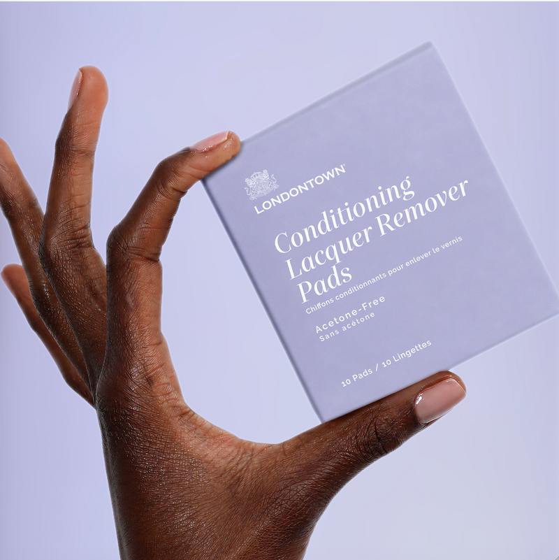 A hand displaying a box of LONDONTOWN Conditioning Lacquer Remover Pads with perfectly manicured nails against a soft lavender background—ideal for easy, on-the-go nail polish removal.