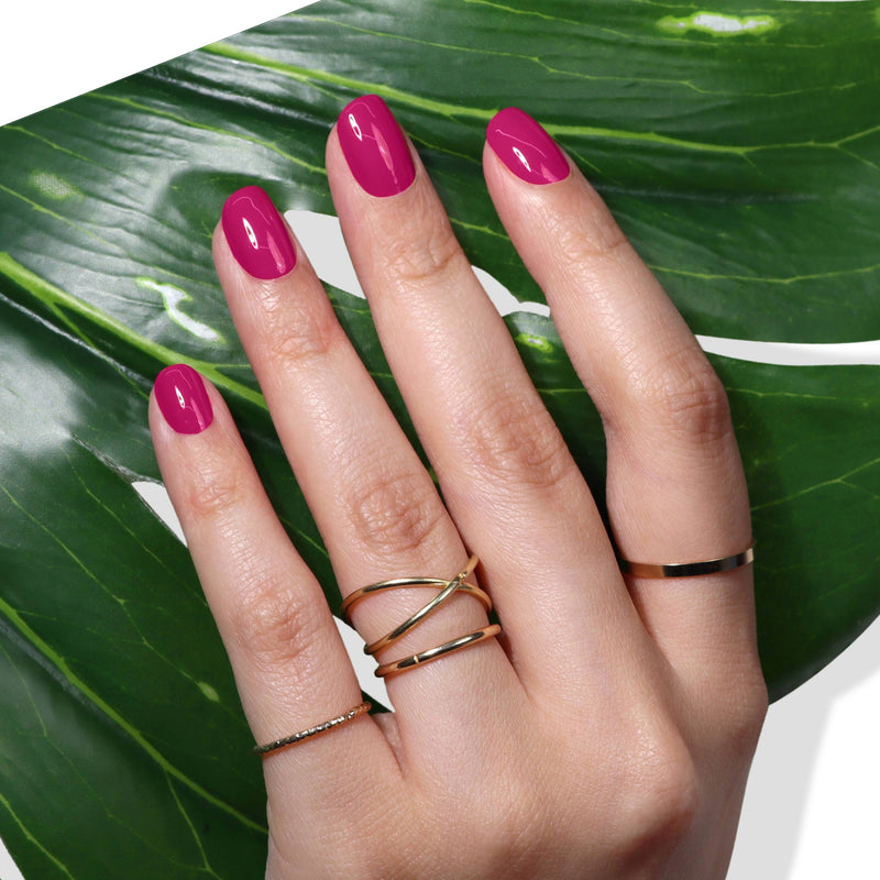 A hand adorned with LONDONTOWN's Teeny 'Kini nail polish and several gold rings rests on a green leaf, evoking a fresh, fruity vibe.