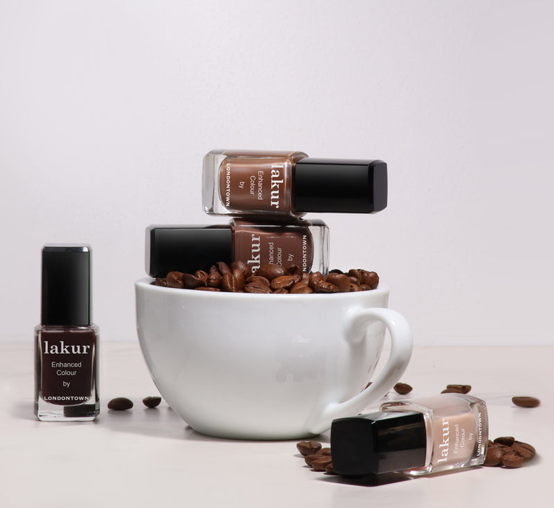 A white coffee mug filled with espresso beans holds three LONDONTOWN Midnight Mocha nail polish bottles, with another bottle next to it on a white surface.